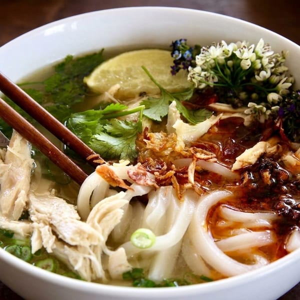 A white bowl of chicken pho with noodles, lime, cilantro, and chili oil, with chopsticks.
