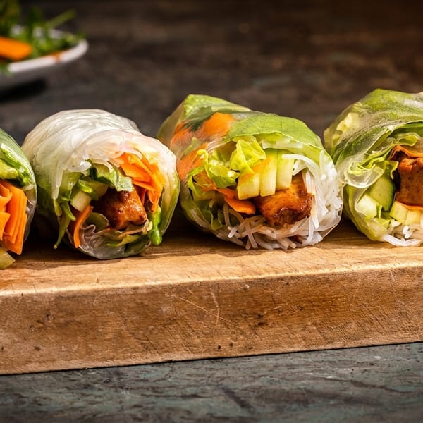 Four fresh spring rolls with tofu, rice noodles, lettuce, and carrots on a wooden board.