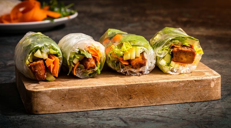 Four fresh spring rolls with tofu, rice noodles, lettuce, and carrots on a wooden board.