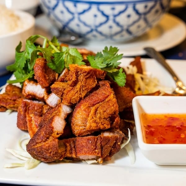 Crispy fried pork belly with cilantro, shredded cabbage, and sweet chili sauce, with rice in the background.