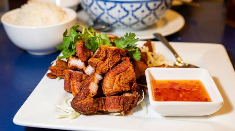 Crispy fried pork belly with cilantro, shredded cabbage, and sweet chili sauce, with rice in the background.