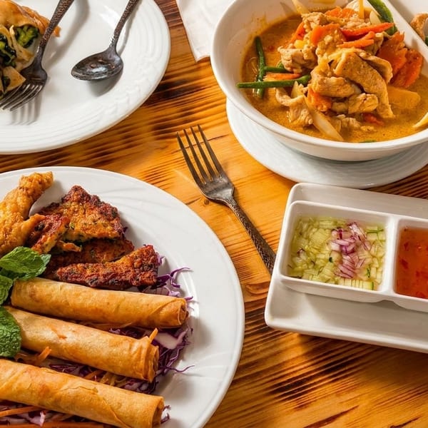 A wooden table laden with various Thai dishes, including curry, stir-fry, spring rolls, and fried chicken.