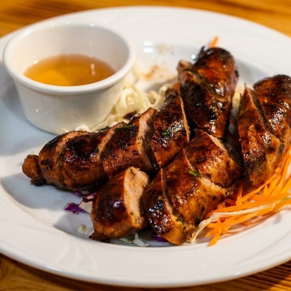 Grilled, sliced sausages with a side of dipping sauce, shredded cabbage, and carrots on a white plate.