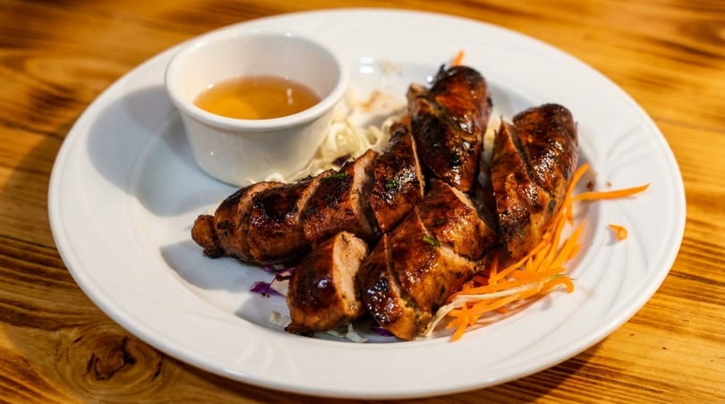 Grilled, sliced sausages with a side of dipping sauce, shredded cabbage, and carrots on a white plate.