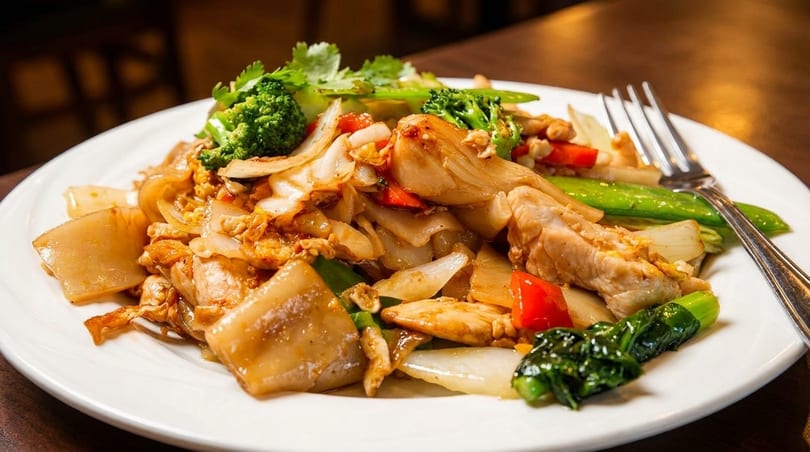 A close-up of a plate of Pad See Ew with chicken, wide noodles, broccoli, and other vegetables.